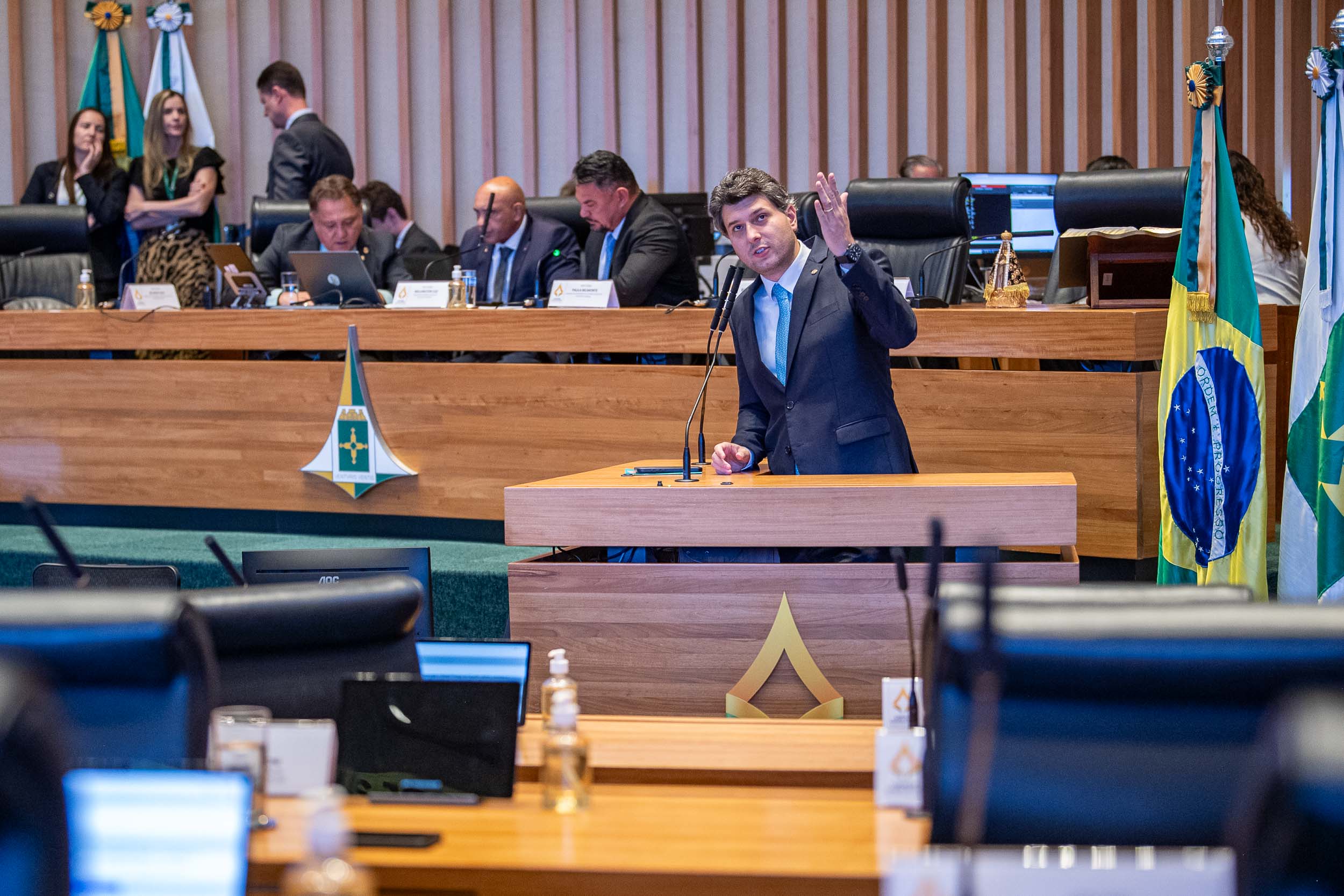 Deputado em pé na Câmara Legislativa do DF com diversos deputados atrás sentados na tribuna
